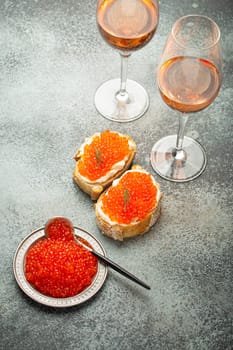Small metal plate with red salmon caviar, two caviar toasts canapé, two glasses of champagne top view on grey concrete background, festive luxury delicacy and appetizer.