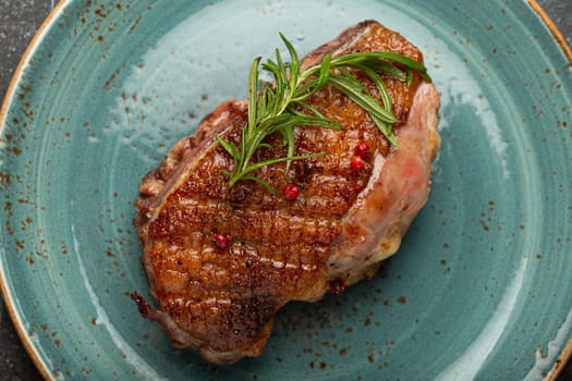 Delicious roasted duck breast fillet with golden crispy skin, with pepper and rosemary, top view on ceramic blue plate, rustic concrete rustic background.