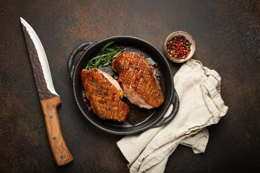 Two roasted duck breast fillets with crispy skin, with pepper and rosemary, top view in black cast iron pan with knife, dark brown concrete rustic background.