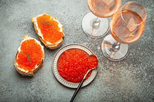 Small metal plate with red salmon caviar, two caviar toasts canapé, two glasses of champagne top view on grey concrete background, festive luxury delicacy and appetizer.