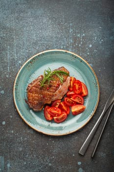 Delicious roasted duck breast fillet with golden crispy skin, with pepper and rosemary, top view on ceramic blue plate served with cherry tomatoes salad, rustic concrete rustic background