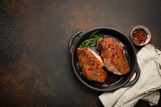 Two roasted duck breast fillets with crispy skin, with pepper and rosemary, top view in black cast iron pan with knife, dark brown concrete rustic background, space for text.