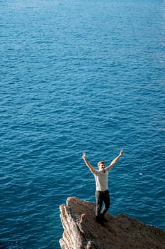 Smiling young man stands on a stone ledge above the sea with his hands up. High quality photo