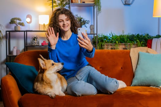 Cheerful young woman with curly hair cute corgi dog, taking selfie via smartphone waving hello sitting on sofa at home living room. Happy girl pet lover spending leisure time with dog during weekend