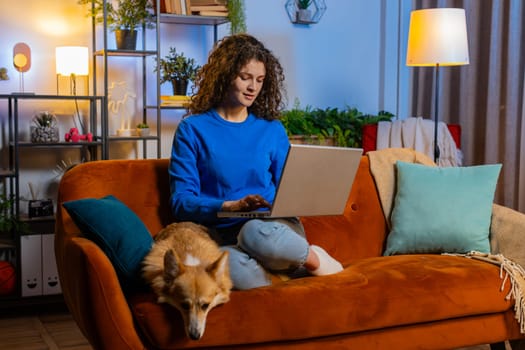 Happy woman with curly hair opening laptop and working remotely sitting beside corgi dog on sofa in living room. Beautiful female girl freelancer in casual clothes smiling and using netbook at home.