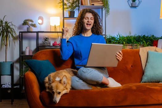 Surprised young woman with curly hair using laptop and celebrating success while sitting on sofa in living room at home. Happy female girl freelancer dancing on online project approval in apartment.