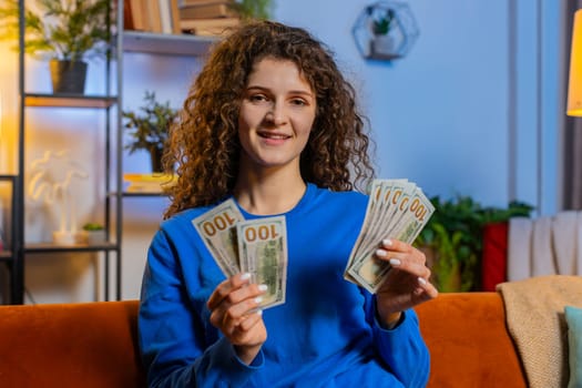 Successful happy rich woman with curly hair counting dollar banknotes while sitting on couch in living room at home. Smiling girl calculating budget, savings, earnings, planning vacation in apartment.