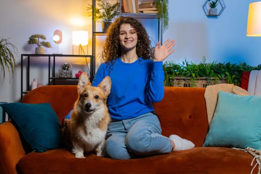 Portrait of smiling young woman with curly hair waving hands saying hello and looking at camera. Cheerful beautiful lady girl in casual clothes greeting while sitting on sofa in living room at home.