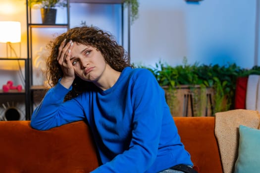 Upset lonely young woman with curly hair in casual clothes thinking about problems sitting on sofa in living room at home. Bored female girl feeling bad annoyed burnout while looking away in apartment