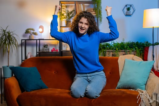 Portrait of happy young woman with curly hair celebrating success showing thumbs up dancing sits on sofa in living room at home. Excited adult girl in casual clothes watching winning match at home.