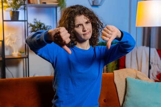 Portrait of upset young woman with curly hair showing thumbs down dislike gesture sitting on couch in living room. Displeased Caucasian female adult girl expressing discontent and disapproval at home