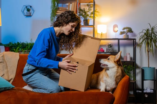 Shocked satisfied woman unpacking delivery parcel. Girl shopper online shop customer opening cardboard box receiving purchase gift by fast postal shipping beside cute corgi dog in living room at home