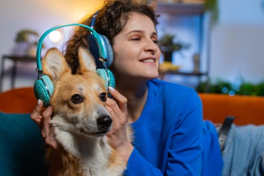 Joyful young woman listening to music through wireless headphones lying on sofa with corgi dog. Happy female girl with curly hair dancing on music in living room at home. Enjoying dance disco music