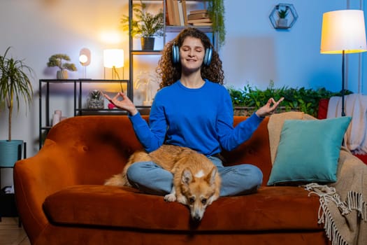 Full body of beautiful young woman with curly hair in headphones meditating in mudra gesture sitting on couch. Relaxed female listening music with eyes closed in living room at home beside corgi dog