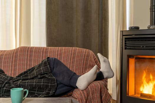 Man's feet with socks near a pellet stove to stay warm, the man is lying on the sofa near the stove to better withstand the cold winter