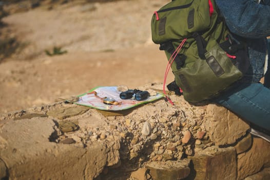 Tourism concept. Close-up binoculars, compass on a map near a backpack of a male tourist sitting on the cliff, enjoying his hiking on the adventure travel.