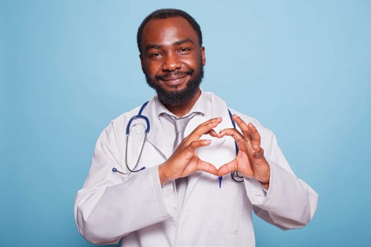 African American medical practitioner with a stethoscope standing over an isolated blue background smiling and showing a heart symbol with hands. Romantic concept.