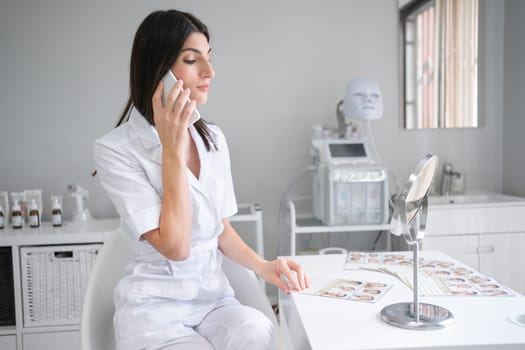 Young female cosmetologist in white uniform talking on smartphone at desk. Woman beautician discussing facial skin treatments for clients on phone call at beauty spa salon.