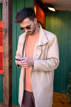 Stylish young man in sunglasses and trench coat text messaging via smartphone. Handsome hipster sending Christmas holiday greetings enjoying festive atmosphere outside wooden house.