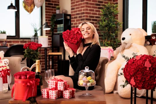 Cute girlfriend holding valentines day red roses bouquet, receiving presents from boyfriend during love holdiday. Woman sitting in living room filled with macarons cookies and romantic presents
