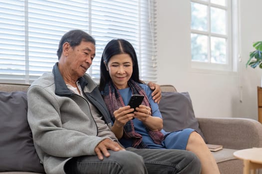 Portrait of Asian adorable senior couple using smartphone together video chatting with family in living room at home..