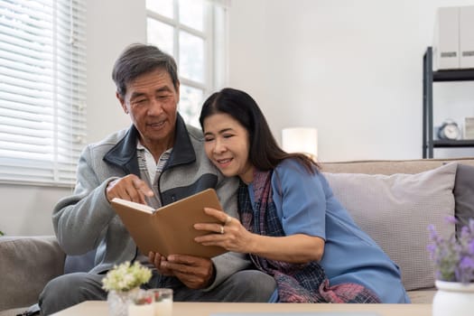 Asian senior couple reading book diary together, sitting on cozy sofa in home interior. Happy retirement activity lifestyle at home.