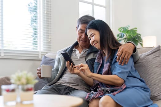 Portrait of Asian adorable senior couple using smartphone together video chatting with family in living room at home..