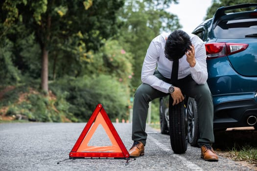 A young businessman in a white shirt confidently changing the wheel of a broken car while a worried mature man sits beside him. A red triangle sign warns drivers of the obstacle ahead.
