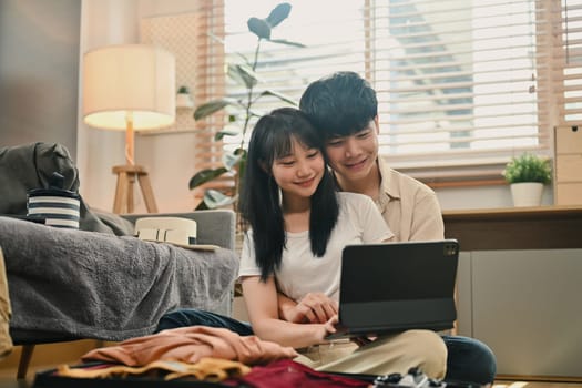 Young couple browsing vacation rental website on digital tablet and packing clothes in living room.