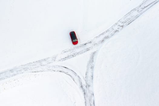 A red car drives down a snowy road with trees and mountains in the background.