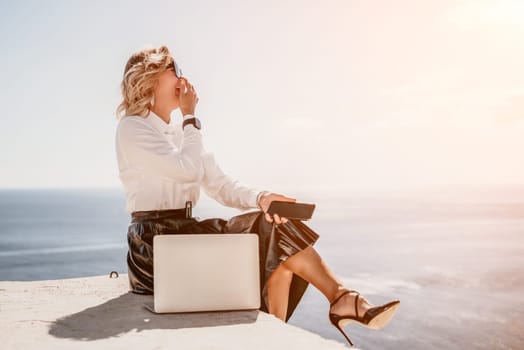 Successful business woman in yellow hat working on laptop by the sea. Pretty lady typing on computer at summer day outdoors. Freelance, travel and holidays concept.