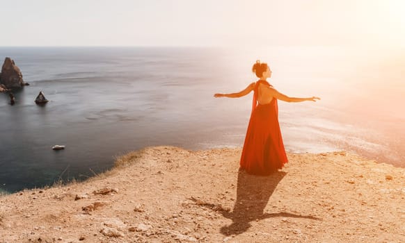 Side view a Young beautiful sensual woman in a red long dress posing on a rock high above the sea during sunrise. Girl on the nature on blue sky background. Fashion photo.