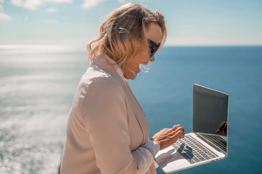 Freelance women sea working on the computer. Good looking middle aged woman typing on a laptop keyboard outdoors with a beautiful sea view. The concept of remote work