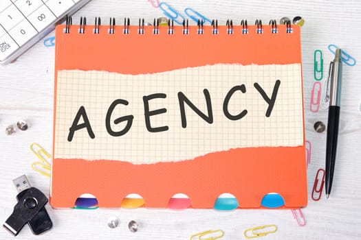 Agency word is written on a sheet in a cage lying on a notebook on the table next to stationery