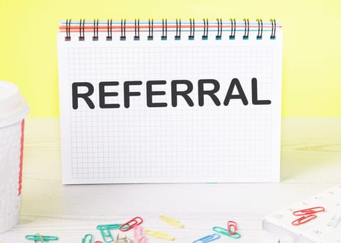 referral the word is written on a blank sheet in a notebook standing on a table on a yellow background