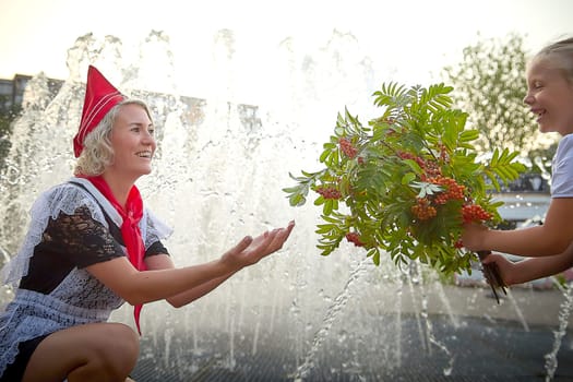 Young and adult schoolgirl on September with flowers having fun near water of fontain. Generations of schoolchildren, pioneer of USSR and October girl in modern uniform of Russia. Mom and daughter