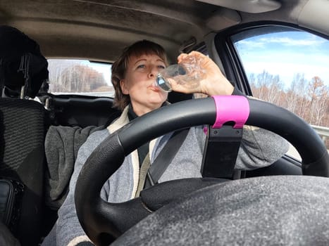 Portrait of female driver in solo journey. Adult mature woman holding steering wheel and bottle of water. Drinking while driving for to stay awake. Lady girl drinks alcohol vodka or gin while driving