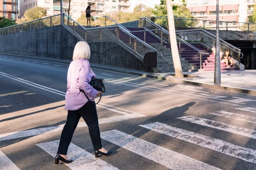 rear view of an unrecognizable senior woman crossing a city street by pedestrian crossing, concept of elderly people leisure and active lifestyle