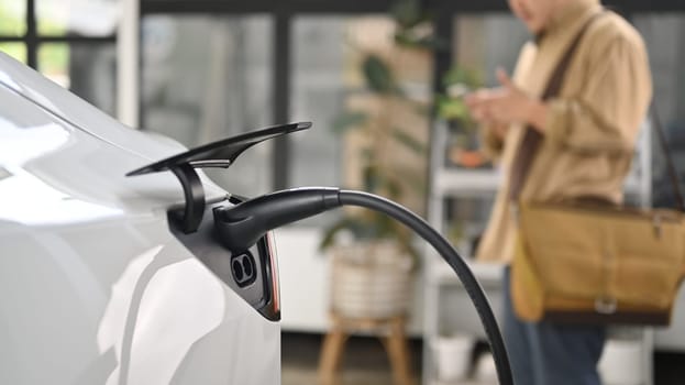 Electric car plugged in to charge outside home with young businessman standing in background.