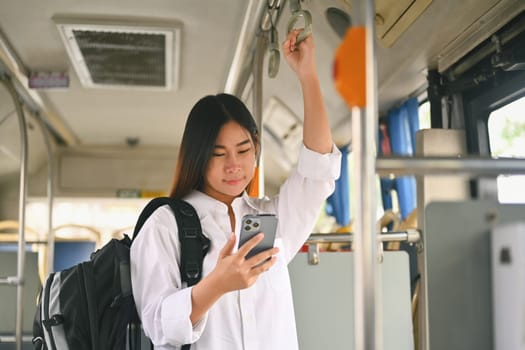 Smiling female traveler with backpack standing in a bus and using mobile phone. Travel and transportation concept.