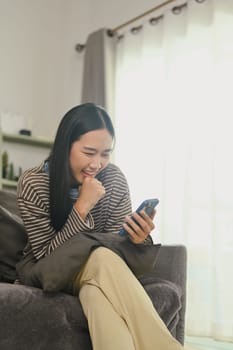 Smiling young woman chatting with friends on mobile phone, sitting on couch at home.