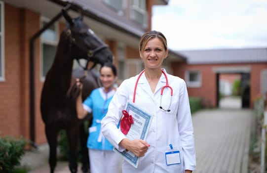 Woman doctor veterinarian holding medical certificate on background of horse. Genetic confirmation of purebred thoroughbred animals concept