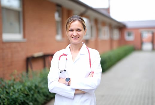 Portrait of young smiling female doctor with red stethoscope near clinic. Professional medical care concept