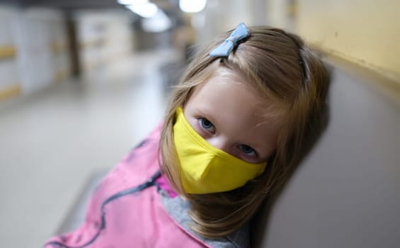 Little girl in yellow protective medical mask sitting in hospital corridor. Medical care for children during covid19 pandemic concept