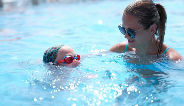 Young woman in sunglasses teaching child to swim in pool. Swimming lessons for children concept