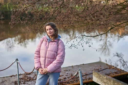 A young woman standing at the shore looking at the river in autumn sunny day. Street view, copy space for text, travel photo. Happy tourist woman on the bank of the river in autumn in warm clothes. Tourists enjoy their vacation, winter season. Romantic look and travel concept. A joyful mood in a Caucasian girl. Winter Wonderland: Enchanting Girl by the Riverside in Autumn.