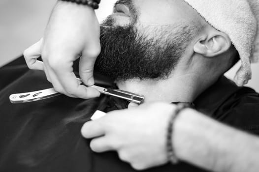 A barber shaves the neck of a bearded customer with a dangerous razor. Shaving the contour of the beard for the correct shape.