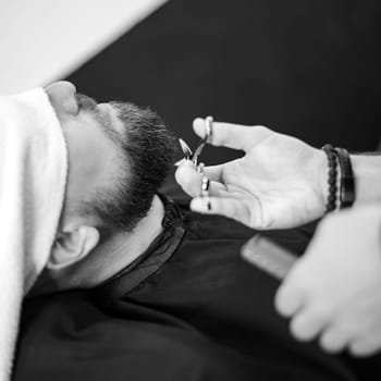 A barber stylist trims the beard of a Caucasian man, whose face is covered with a towel, with scissors