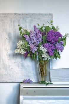 a bouquet of lilacs in a transparent vase on a light textured background. vertical image of lilac, violet and white flowers. spring or summer card. soft focus