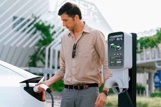 Young man put EV charger to recharge electric car's battery from charging station in city commercial parking lot. Rechargeable EV car for sustainable environmental friendly urban travel. Expedient